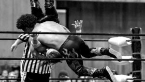 Two wrestlers flying through the air with a referee behind them in a ring with a crowd watching
